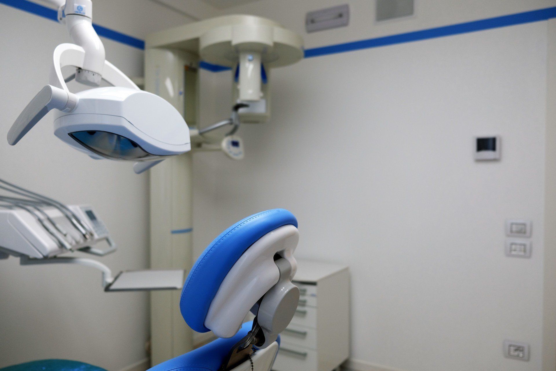 Poltrona odontoiatrica con poggiatesta blu, lampada da soffitto e macchina per raggi X in una stanza bianca.