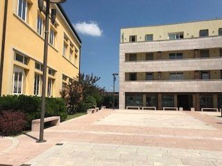 Complesso di due edifici con una piazza pavimentata, una gialla e una beige, contro un cielo blu.