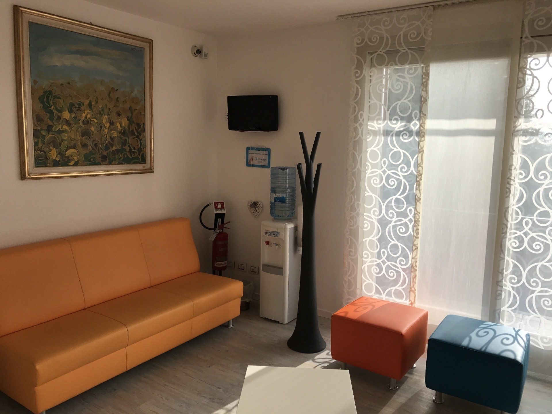 Sala d'attesa: divano arancione, piccola TV, refrigeratore d'acqua, attaccapanni, opere d'arte, finestra con tende a fantasia.
