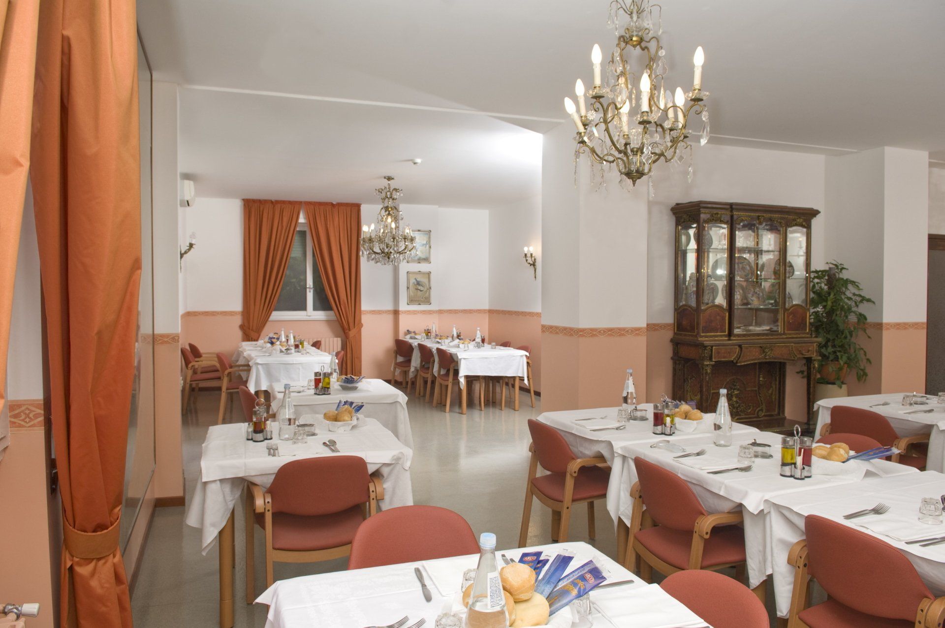 Elegante sala da pranzo con tovaglie bianche, sedie rosse, lampadari e tende arancioni.