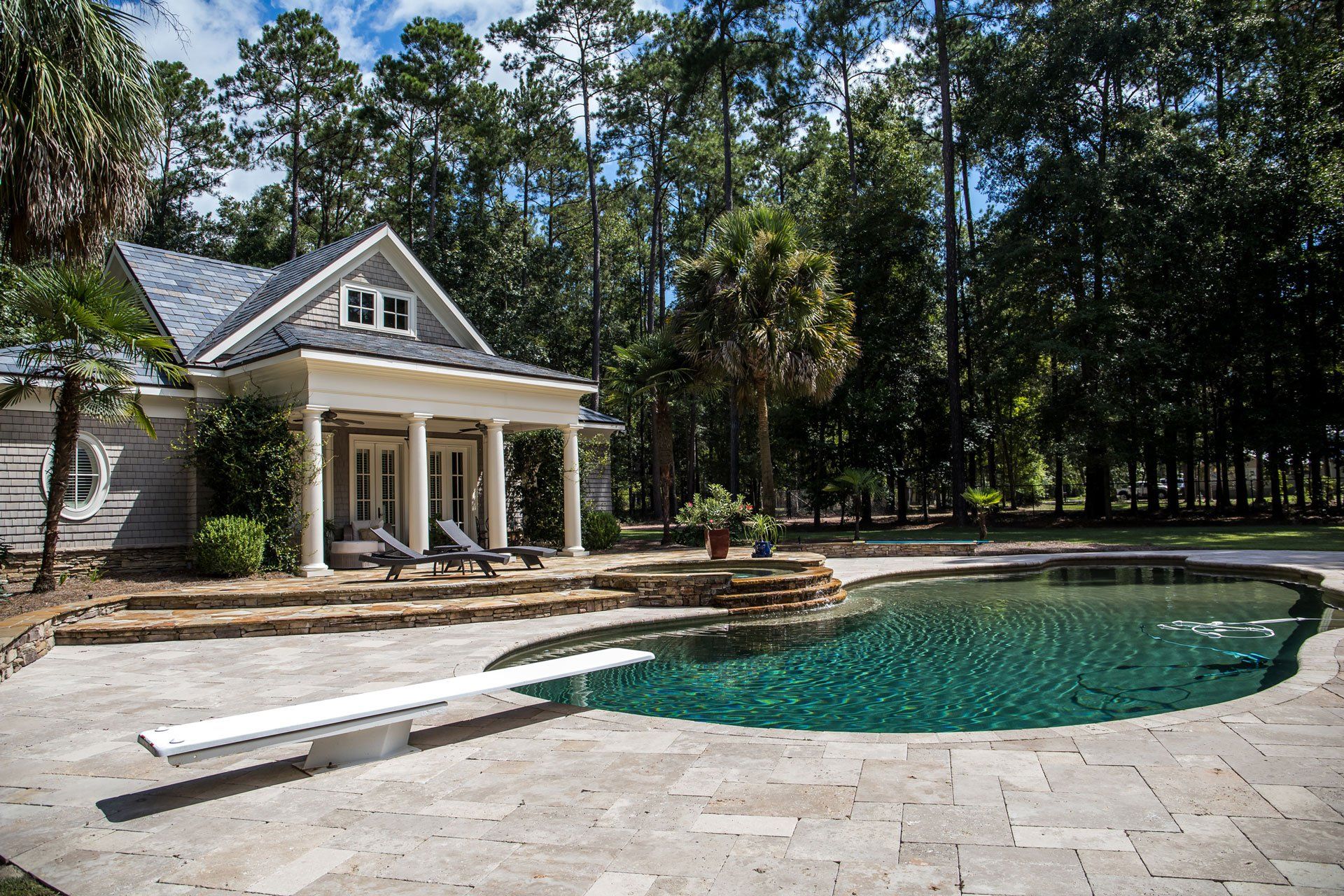 A pool with a diving board sits on a stone patio before a house with a porch, surrounded by trees.