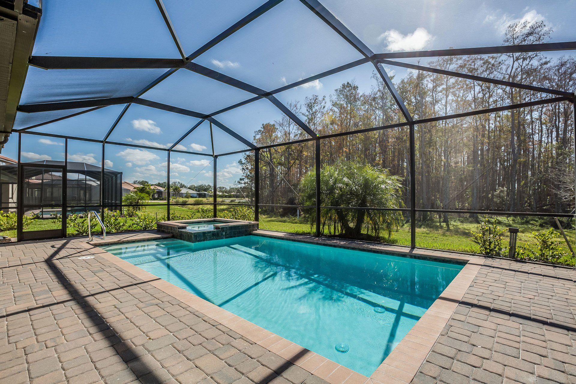 A swimming pool with a spillover spa inside a screened lanai, overlooking a grassy area and trees on a sunny day.