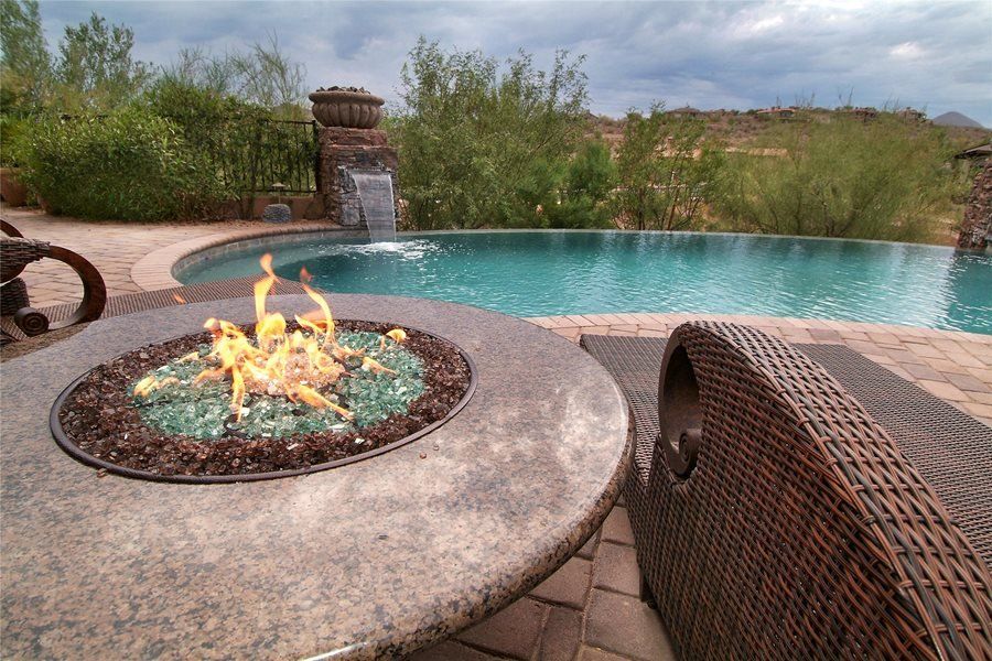 A stone fire pit with glowing green glass rocks overlooks a resort-style swimming pool with a waterfall in the desert.