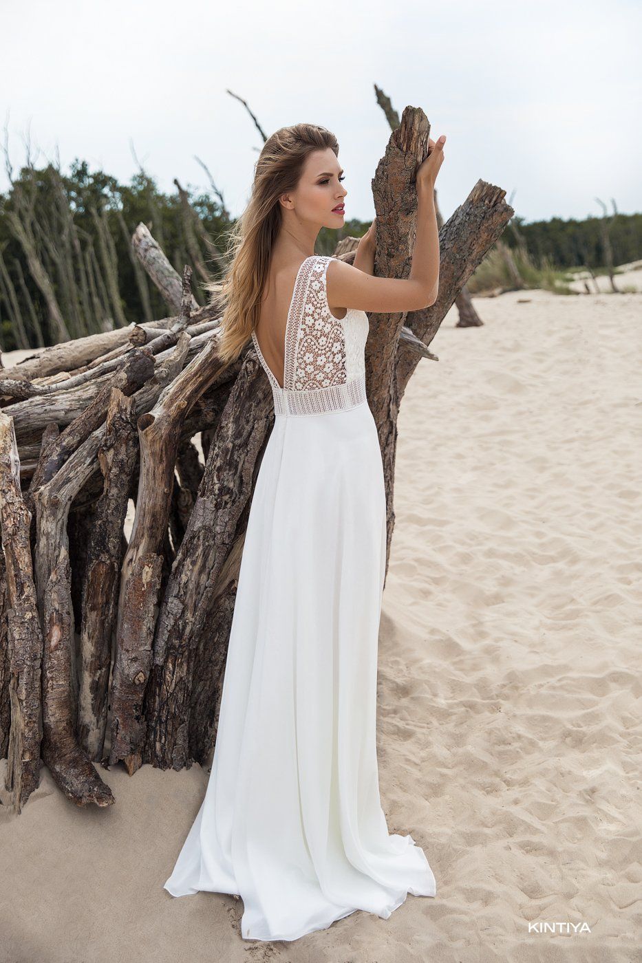 Eine Frau in einem weißen Brautkleid steht neben einem Stapel Baumstämme am Strand.
