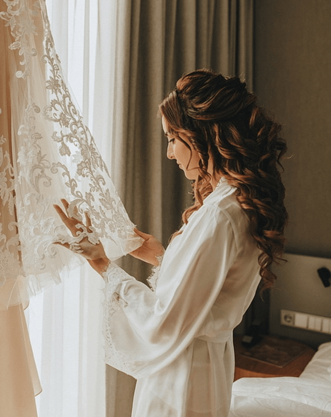 Eine Frau in einem weißen Gewand betrachtet ihr Hochzeitskleid, das an einem Fenster hängt.