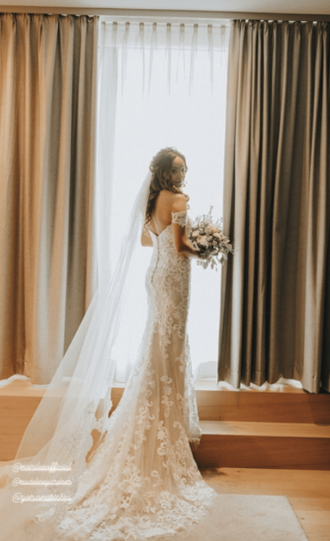 Eine Braut im Brautkleid steht mit einem Blumenstrauß in der Hand vor einem Fenster.