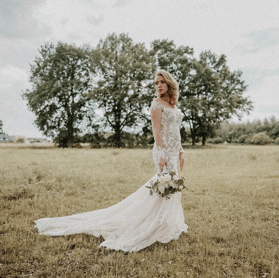 Eine Frau in einem Brautkleid steht mit einem Blumenstrauß in der Hand auf einem Feld.