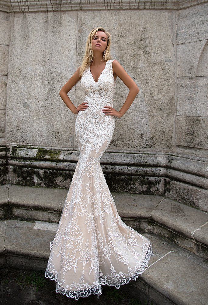 Eine Frau in einem weißen Spitzenbrautkleid steht vor einer Steinmauer.