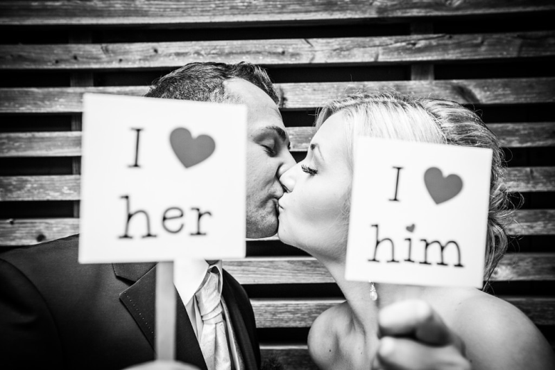 Eine Braut und ein Bräutigam küssen sich und halten Schilder mit der Aufschrift „Ich liebe sie“ und „Ich liebe ihn“ hoch.