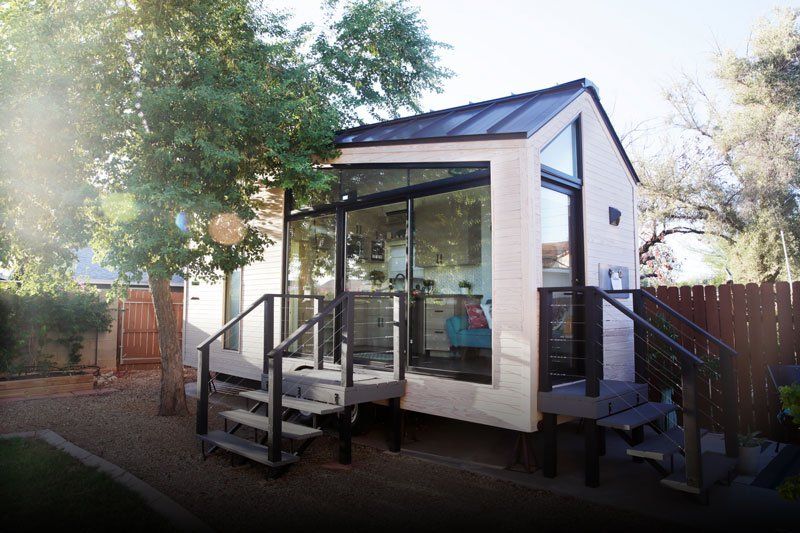 A tiny house with large glass windows, sits in the backyard surrounded by a wooden fence and trees.