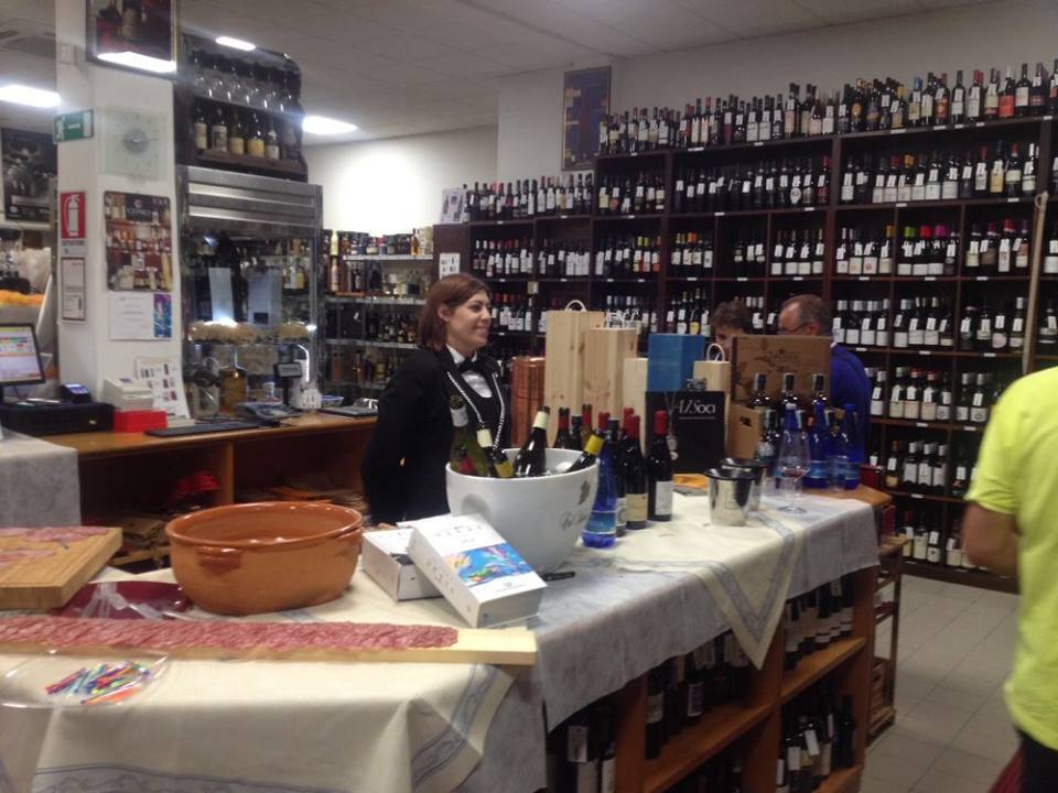 In un'enoteca, una persona sorridente è in piedi dietro un tavolo, con scaffali pieni di bottiglie di vino che ricoprono le pareti.