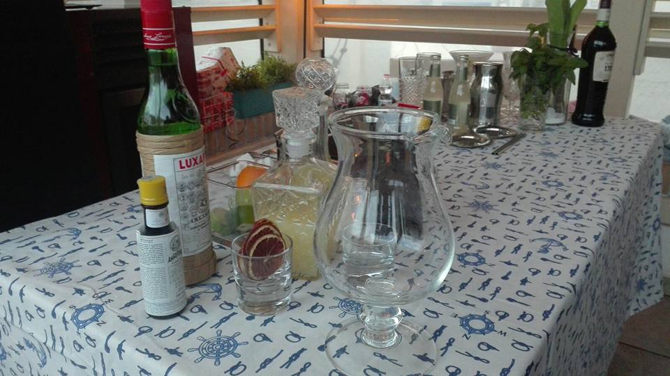 Su un tavolo ricoperto da una tovaglia con stampa nautica sono disposte bottiglie di liquori, un bicchiere da miscelazione, guarnizioni e accessori per cocktail.