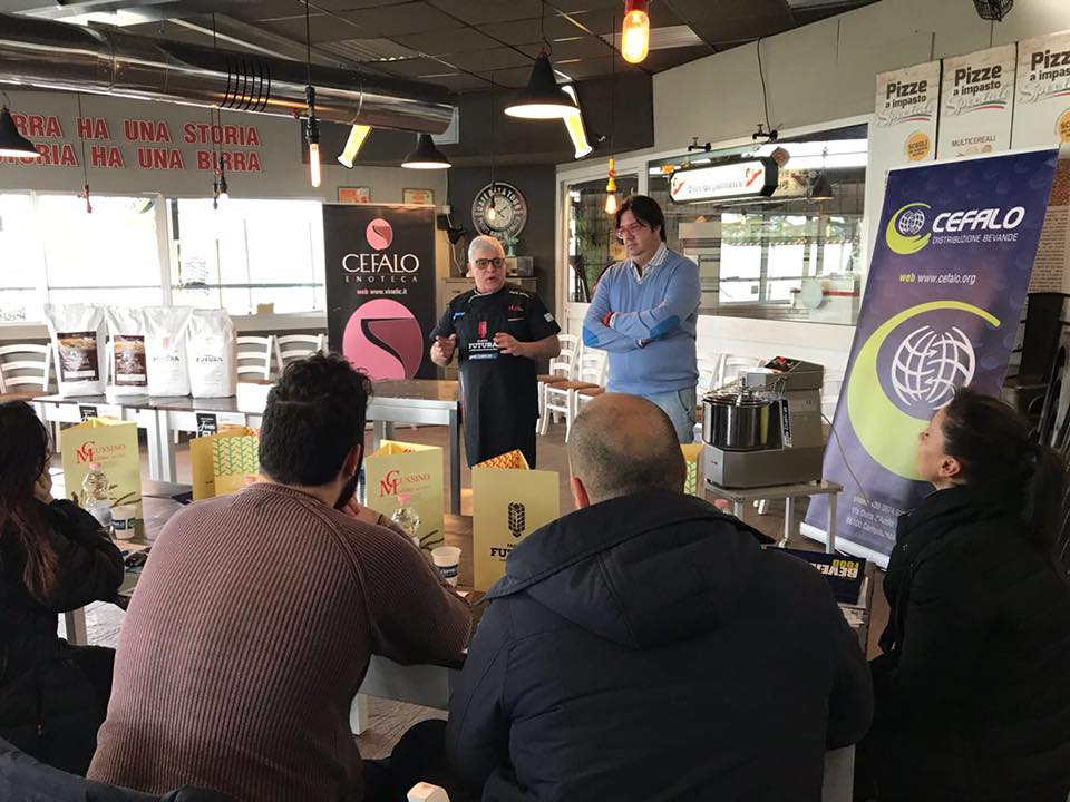 Un relatore si rivolge a un piccolo gruppo di partecipanti durante un workshop al chiuso, incentrato su insegne personalizzate e prodotti a base di farina.