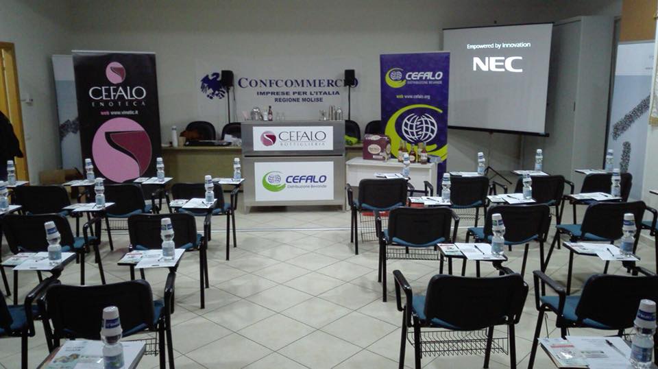 Una sala conferenze vuota con file di sedie, scrivanie con bottiglie d'acqua, striscioni con il logo aziendale e uno schermo per le proiezioni.