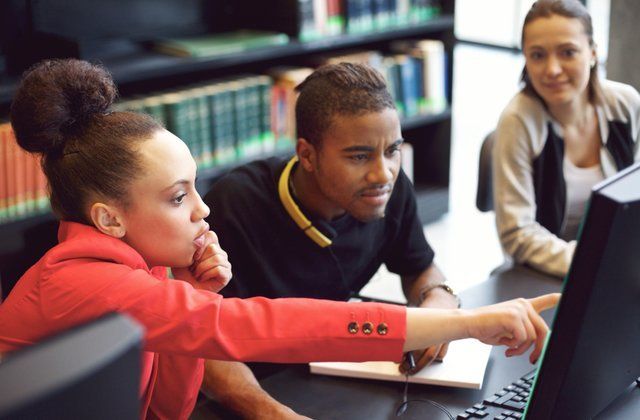 Three people collaborating at a computer in a library. A woman points at the screen, another looks on.