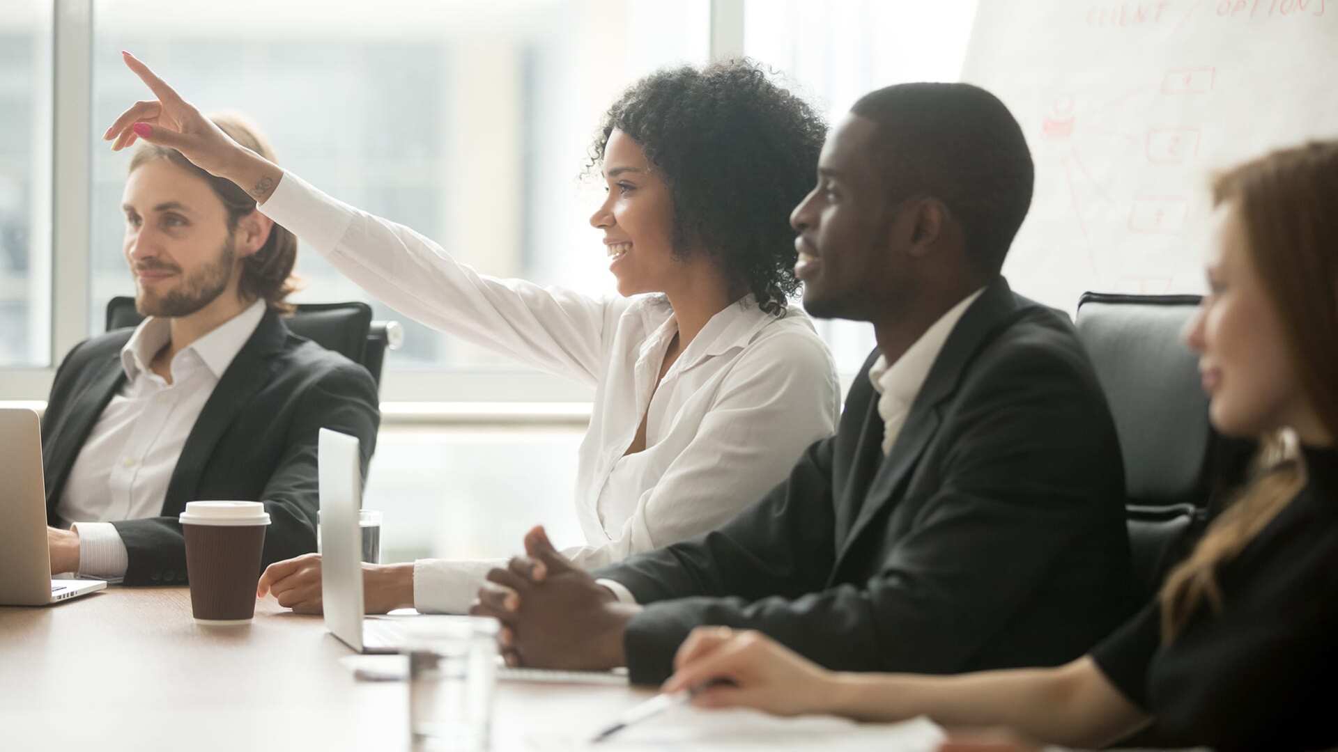 Business people in a meeting, one raising their hand with a question.
