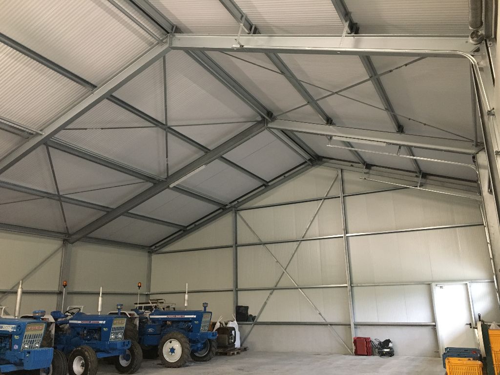 Een grote, lege landbouwschuur met een metalen frame, lichte wanden en blauwe tractoren die ernaast geparkeerd staan.