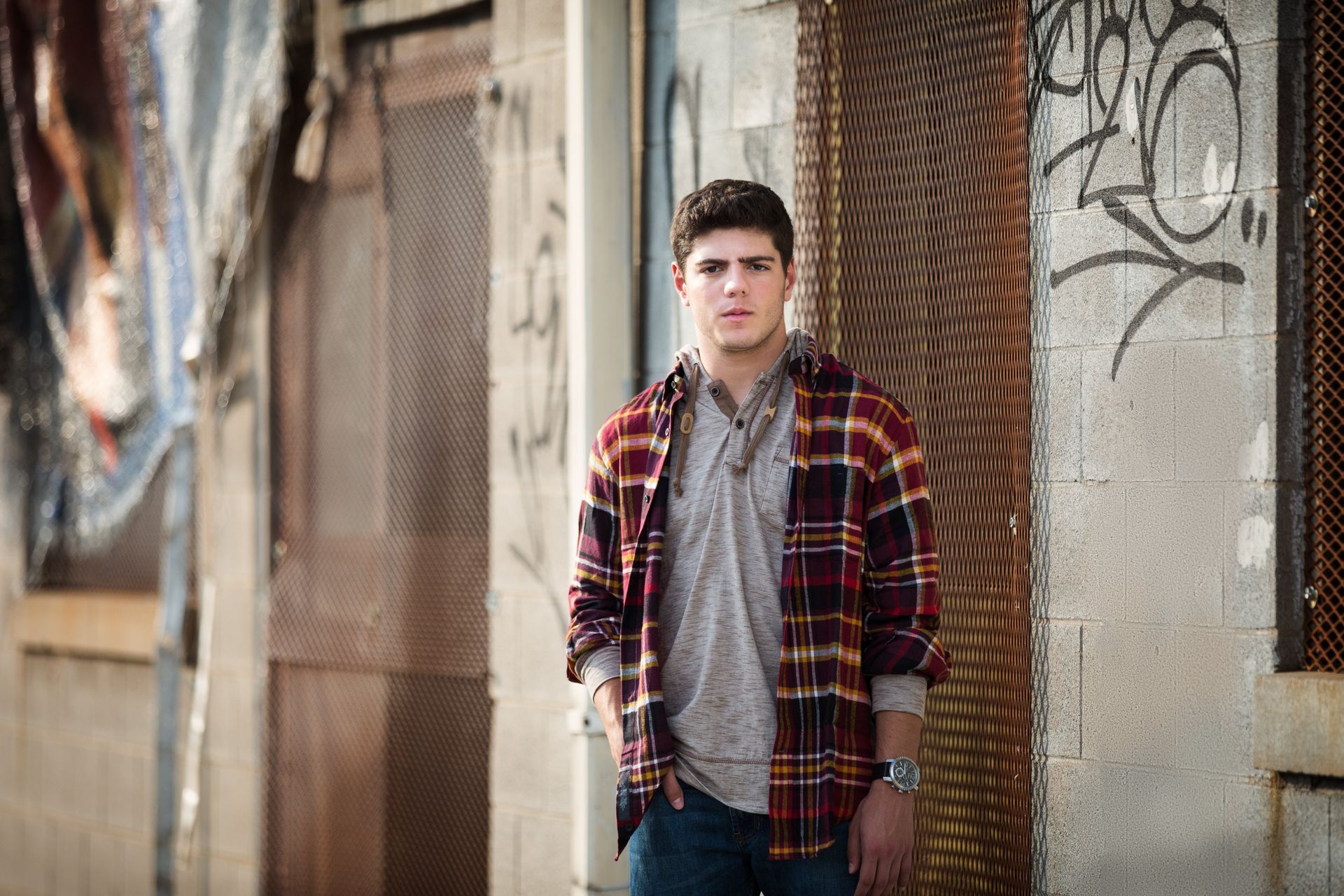 A young man in a plaid shirt is standing in front of a building with graffiti on it.