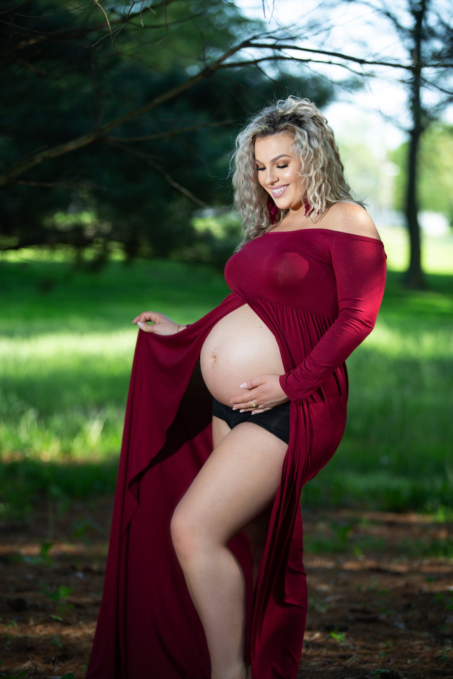 A pregnant woman in a red dress is standing in a park.