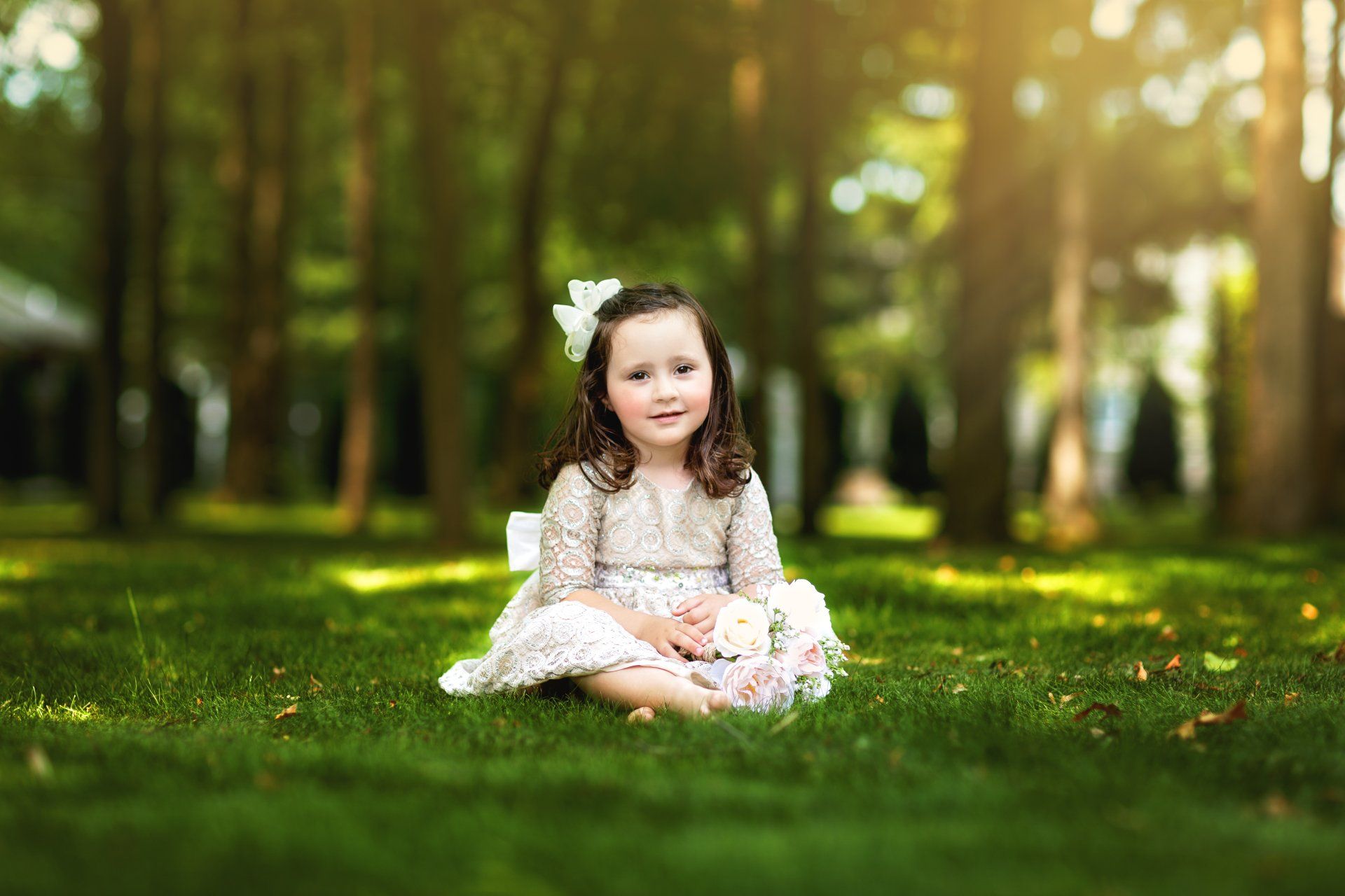 A little girl in a white dress is sitting on the grass in a park.