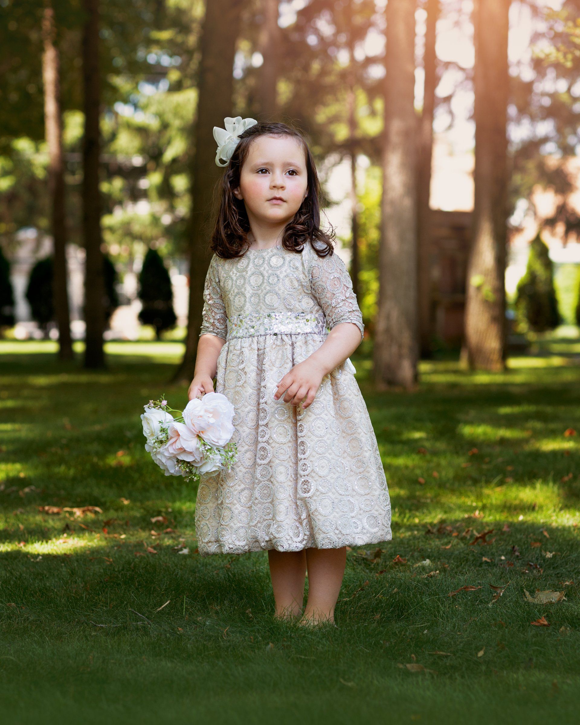 A little girl in a white dress is standing in the grass holding a bouquet of flowers.