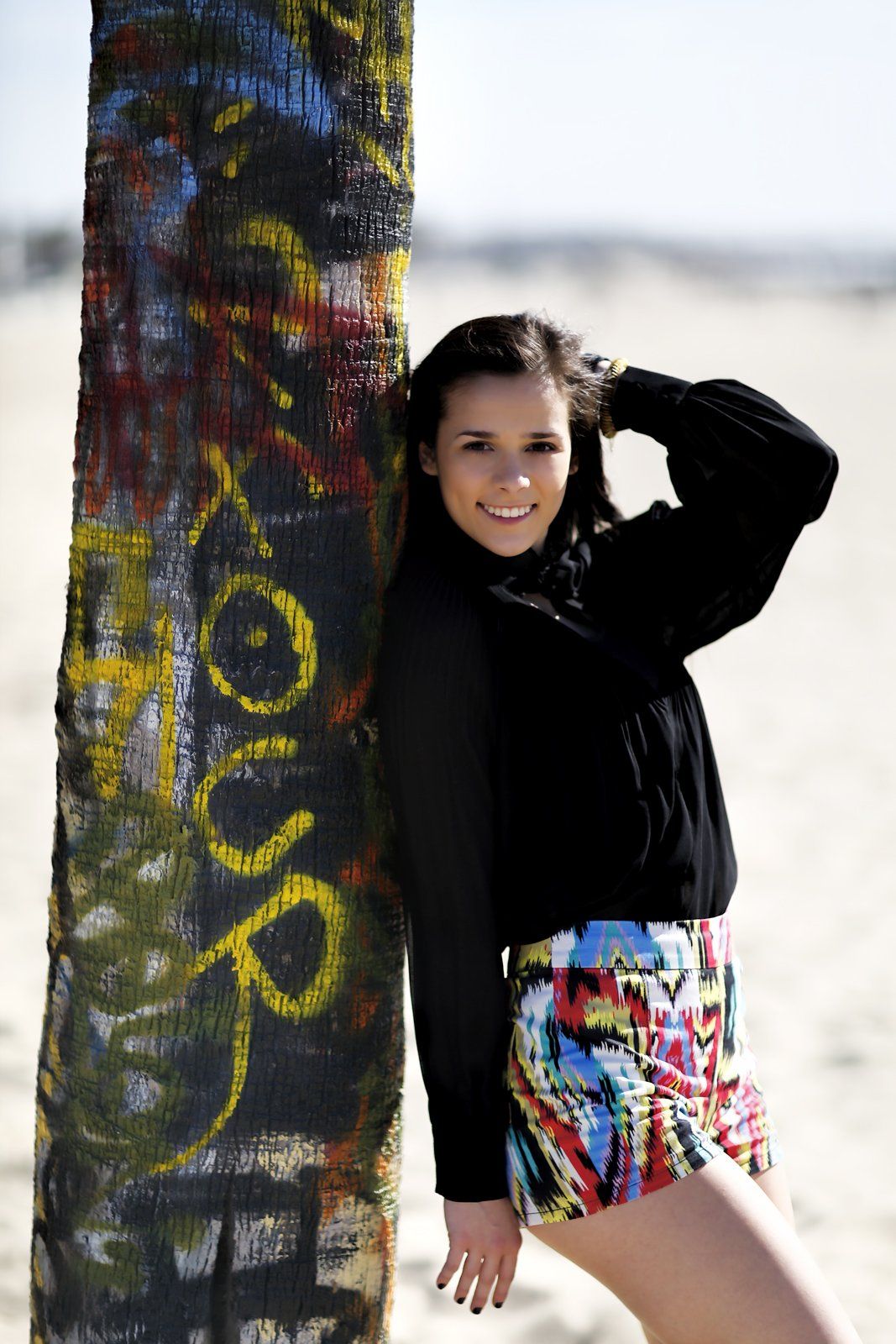 A woman leans against a graffiti covered pole on the beach