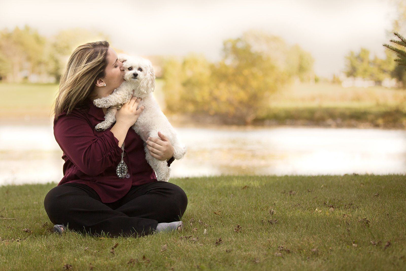 A woman is sitting on the grass kissing a small white dog.