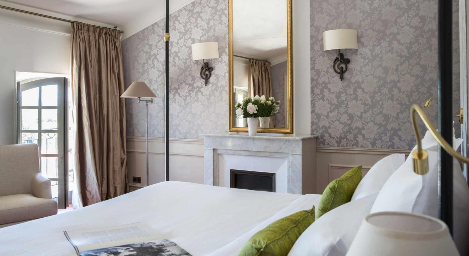 Une chambre avec un grand lit, cheminée, miroir et lampe.