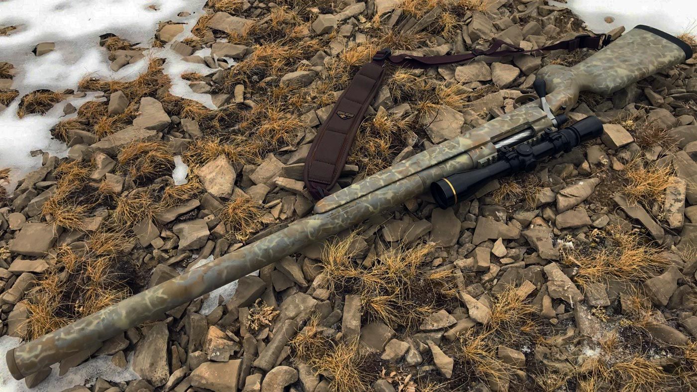 Camouflage-patterned rifle with a scope and strap, lying on rocky ground with dry vegetation and patches of snow.