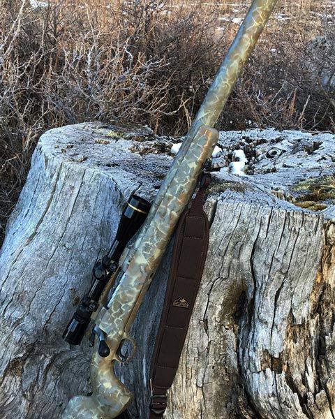 Camouflage-patterned rifle with scope and strap resting on a weathered tree stump outdoors.