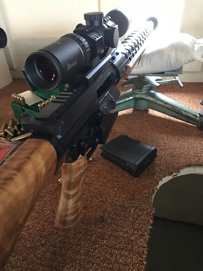 Black rifle with scope, wood grip, and magazine on brown carpet.