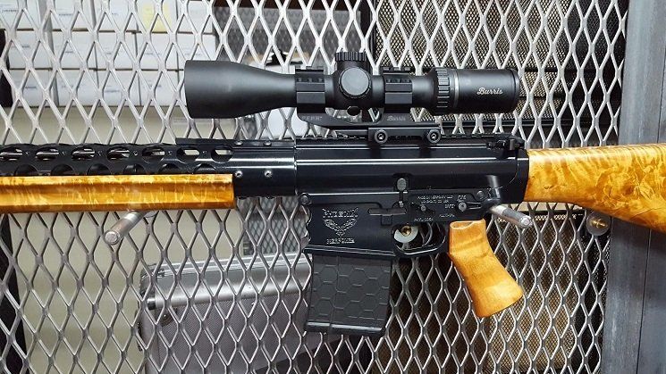 Black and wood-toned rifle with a scope, against a chain link backdrop.