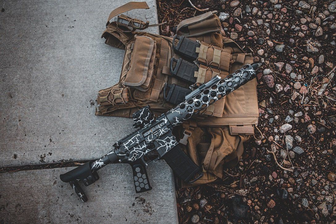 Tactical vest and camouflage-patterned rifle on concrete and gravel ground.