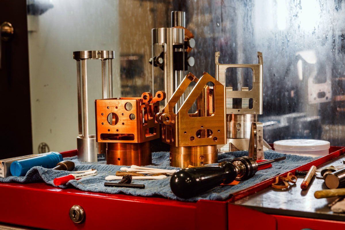 Machined metal parts, copper and silver, on a red workbench with tools. Shop background, tools, and a window.