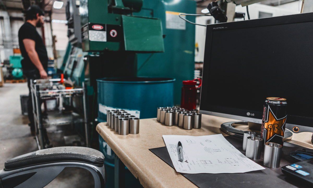 Factory scene: Desk with metal cylinders, monitor, and paperwork. A person stands near machinery.