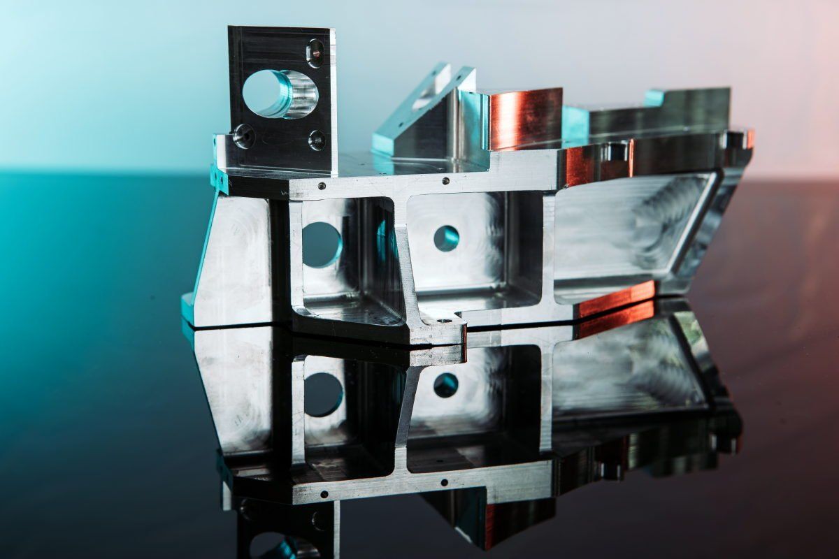 Silver metal mechanical part with several openings, sitting on a reflective surface.