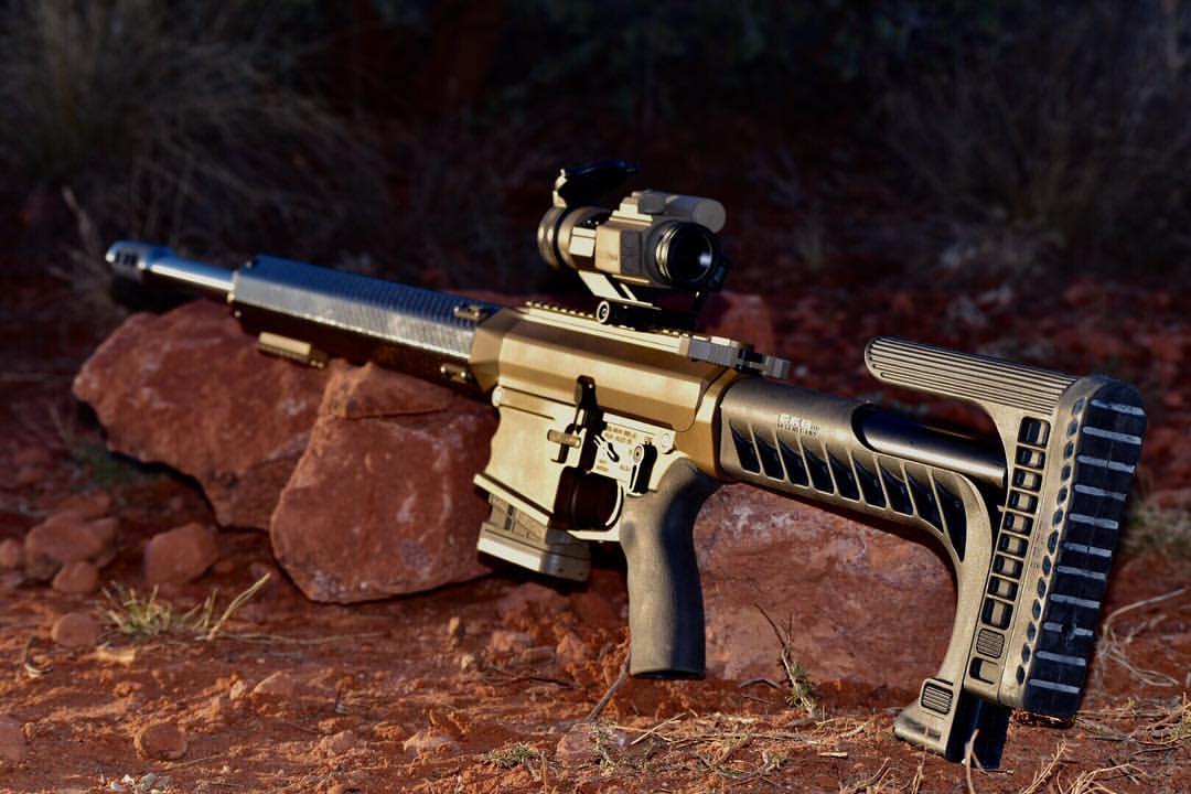 Tan and black AR-style rifle with red dot sight, resting on a brown rock.