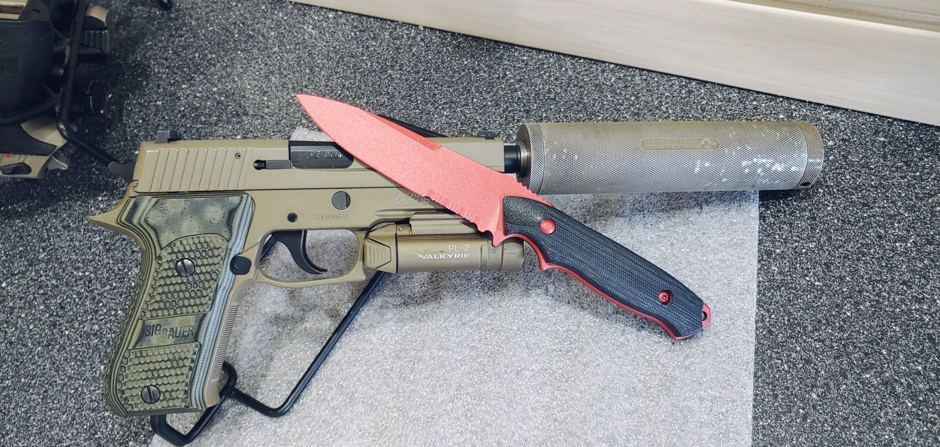 A handgun with a suppressor and a red-bladed knife on a speckled surface.
