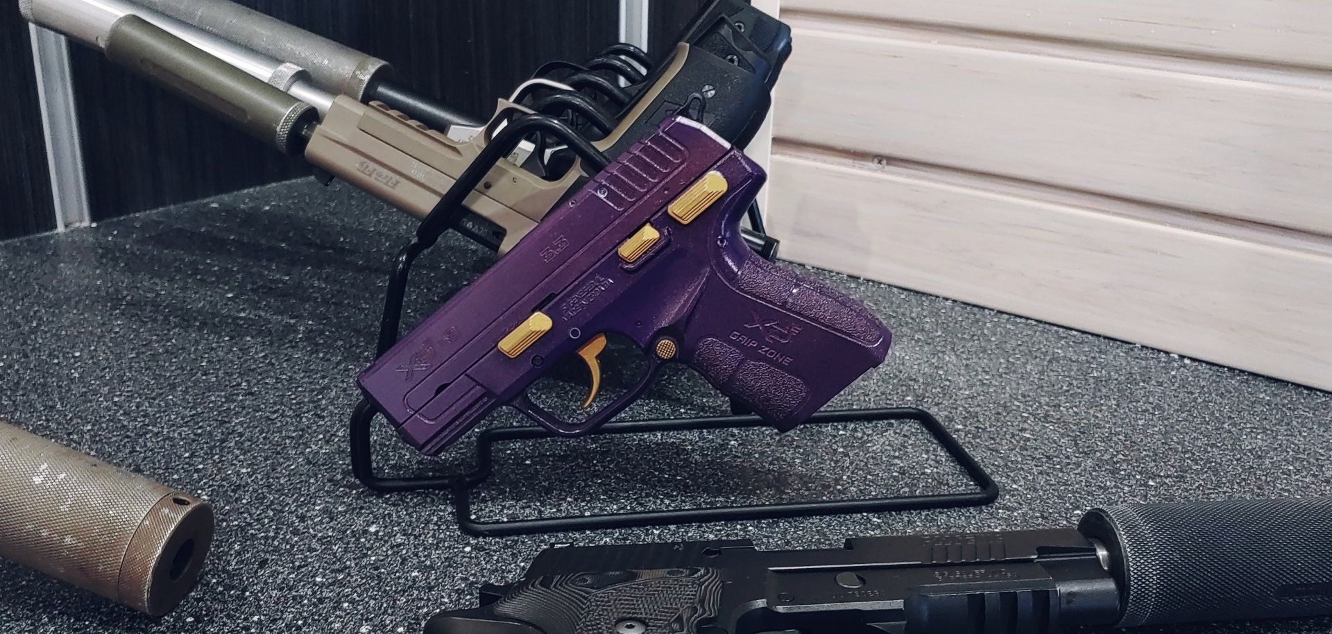 A purple handgun with gold accents displayed on a black stand. Other firearms are also present.