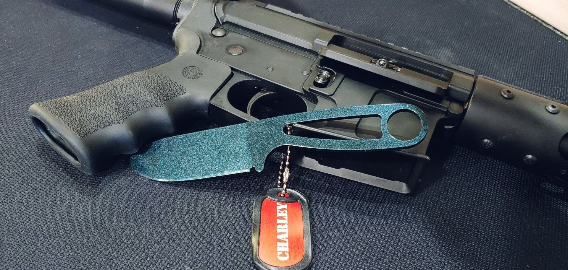 Black firearm with a blue knife and dog tag on a dark surface.