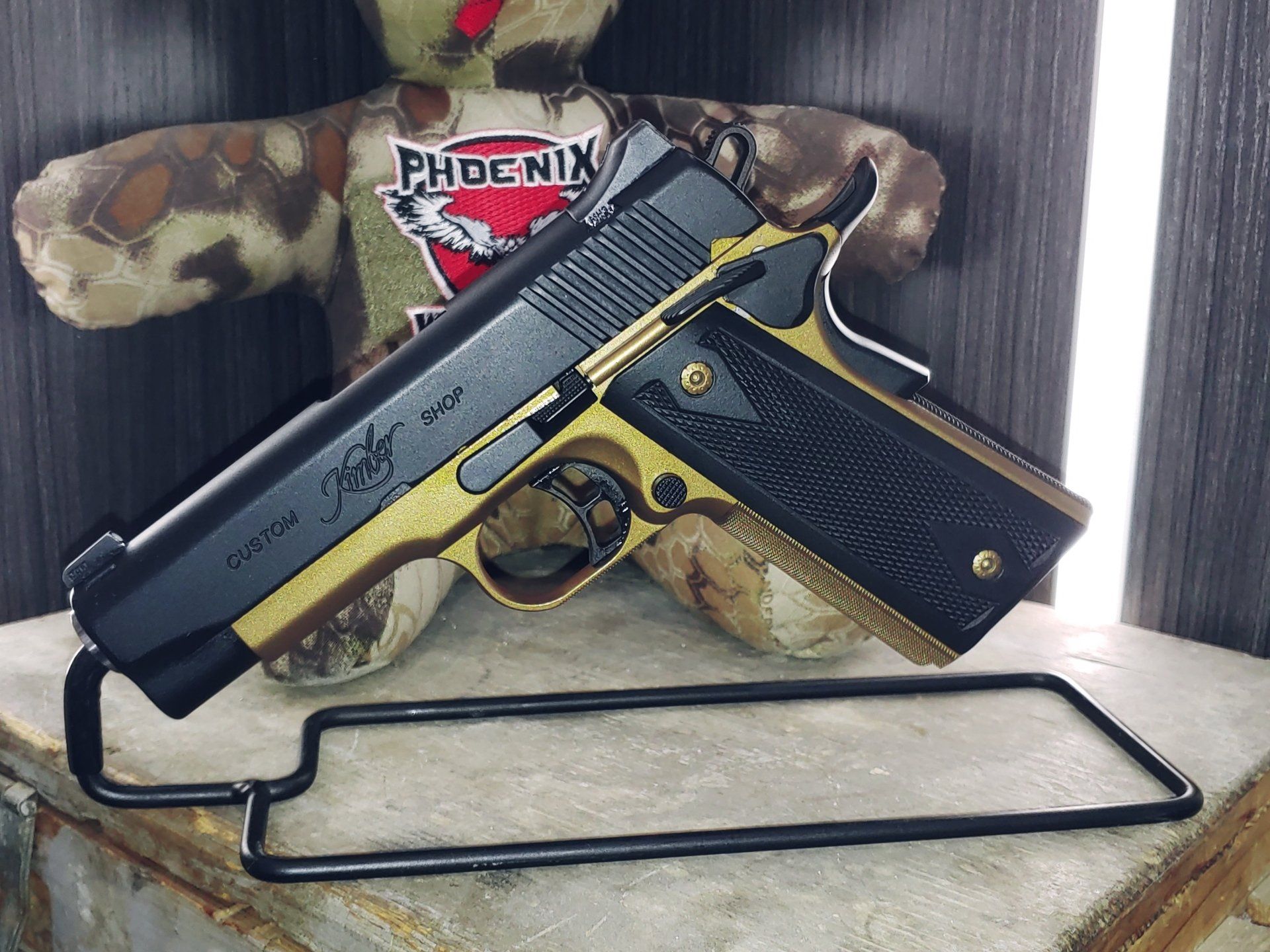 Black and gold handgun on a stand, with a camouflage teddy bear behind it.