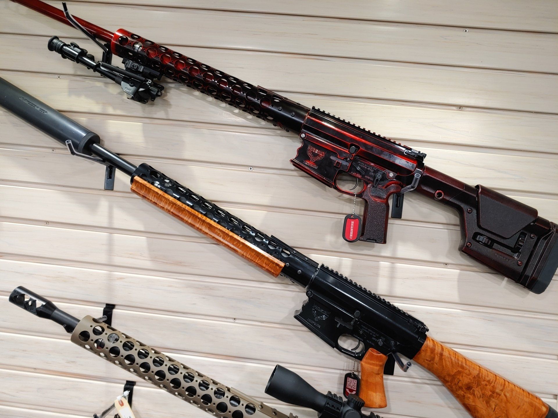 Three rifles displayed on a wall. One red and black, one black and wood, and one with a tan camouflage pattern.