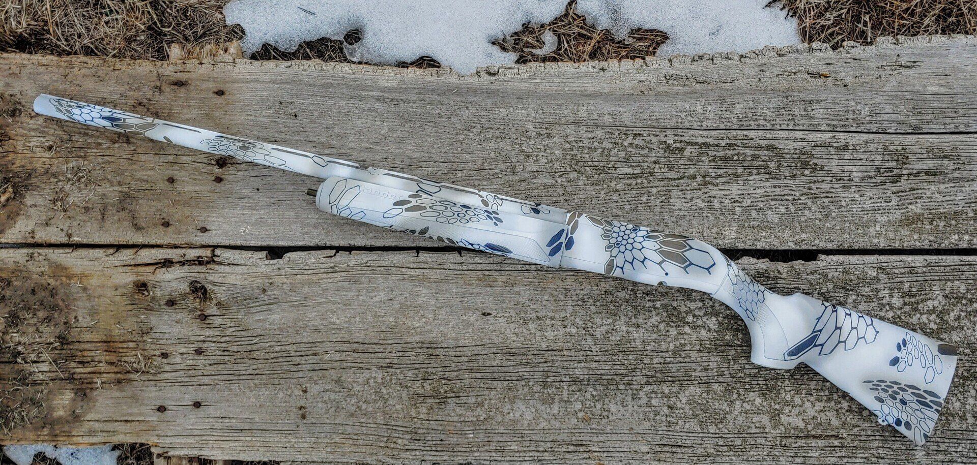 A rifle with a white and blue camouflage pattern rests on a weathered wooden surface with snow.