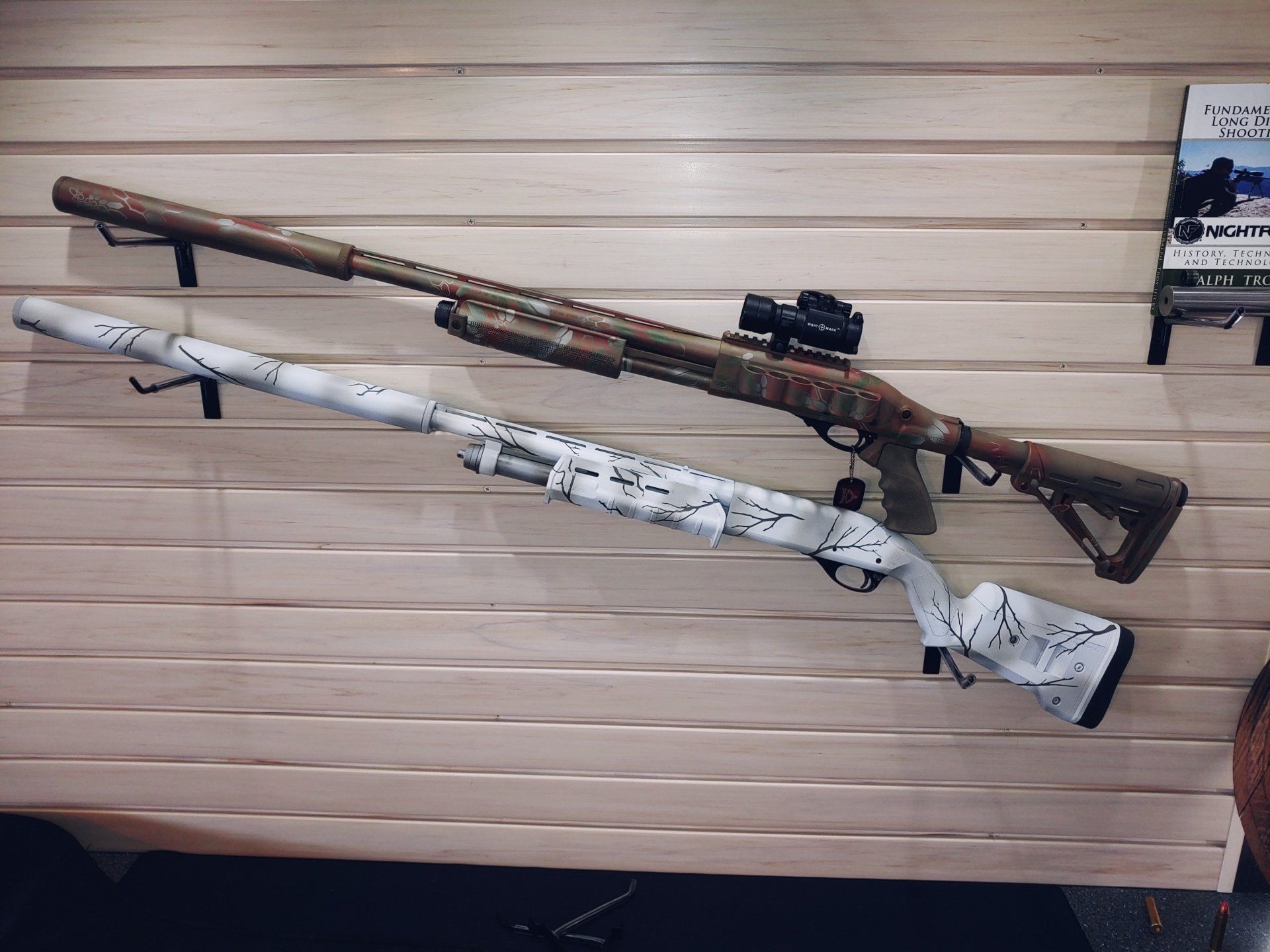 Two camouflage shotguns mounted on a wood-paneled wall. One is brown, the other white.