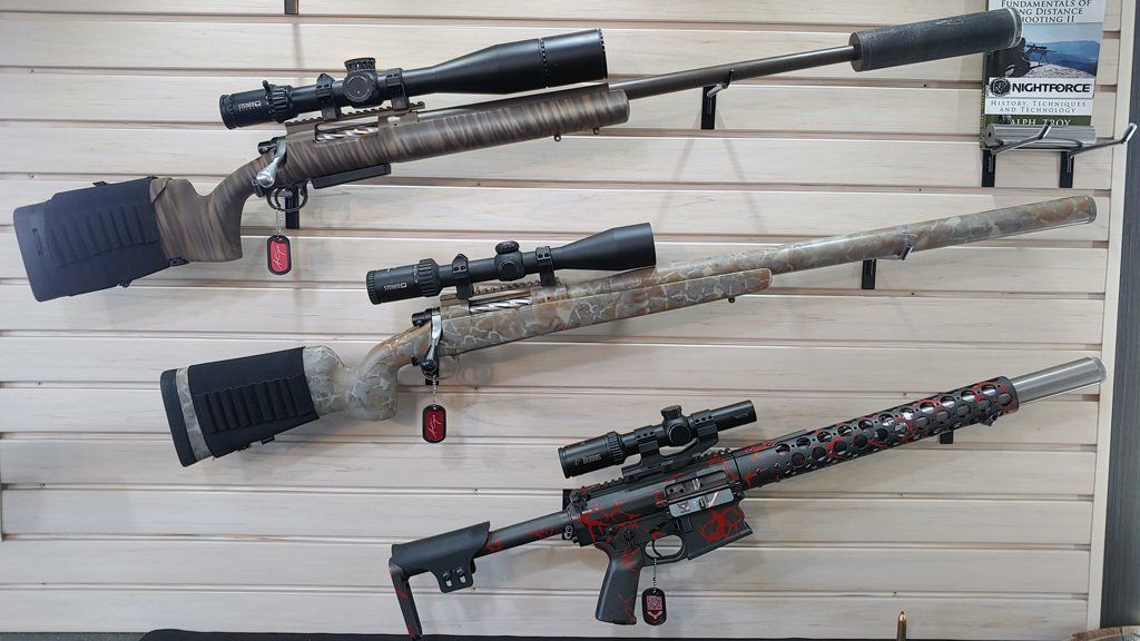 Three rifles with scopes and suppressors mounted on a wooden wall for display.