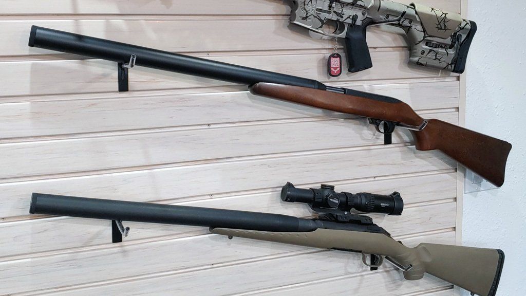 Rifles mounted on a wall. One is camouflaged, the others are black and brown. One has a scope.