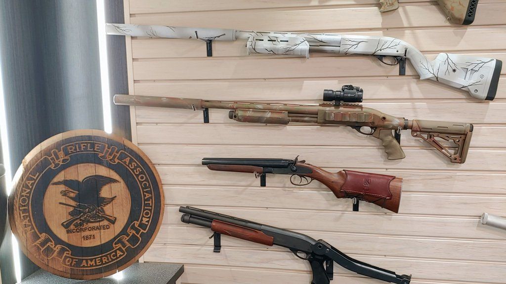 Shotguns mounted on a wooden wall, including a camo-painted one with a scope. A wooden plaque is in the left foreground.