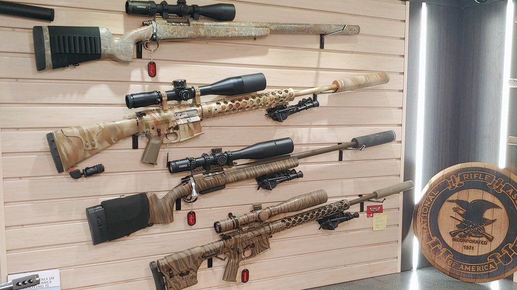 Rifles with scopes on display, mounted on a wooden wall. Camouflage patterns and black accessories are visible.