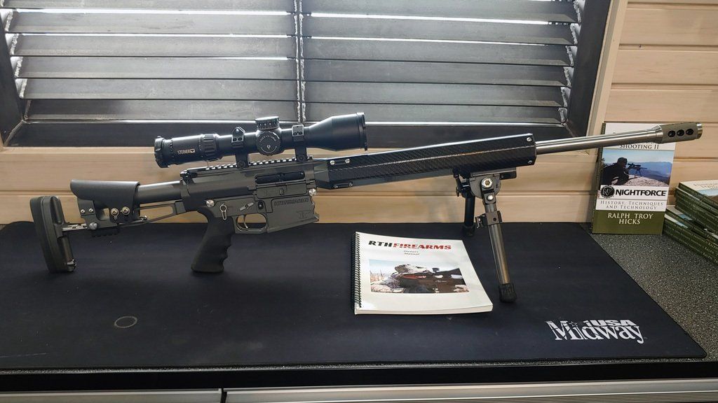 Black rifle with scope and bipod on a mat, magazine in front, indoors.