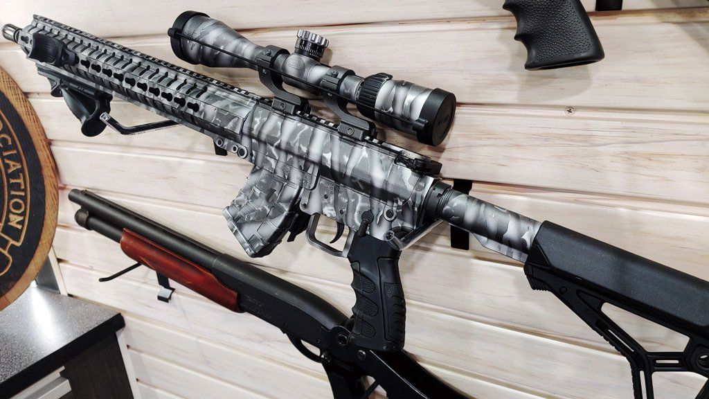 Camouflage rifle with scope and black shotgun mounted on wooden wall.